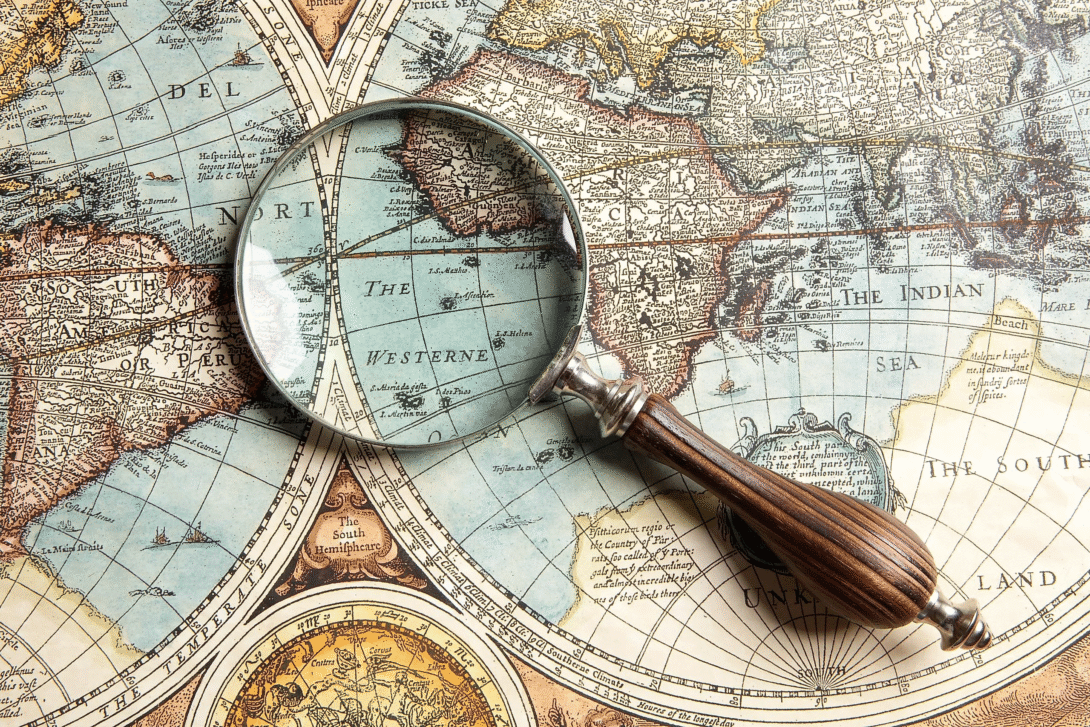 A magnifying glass reveals the intricate hand-drawn detail of an antique world map — cartography's enduring art of translating the Earth onto paper.